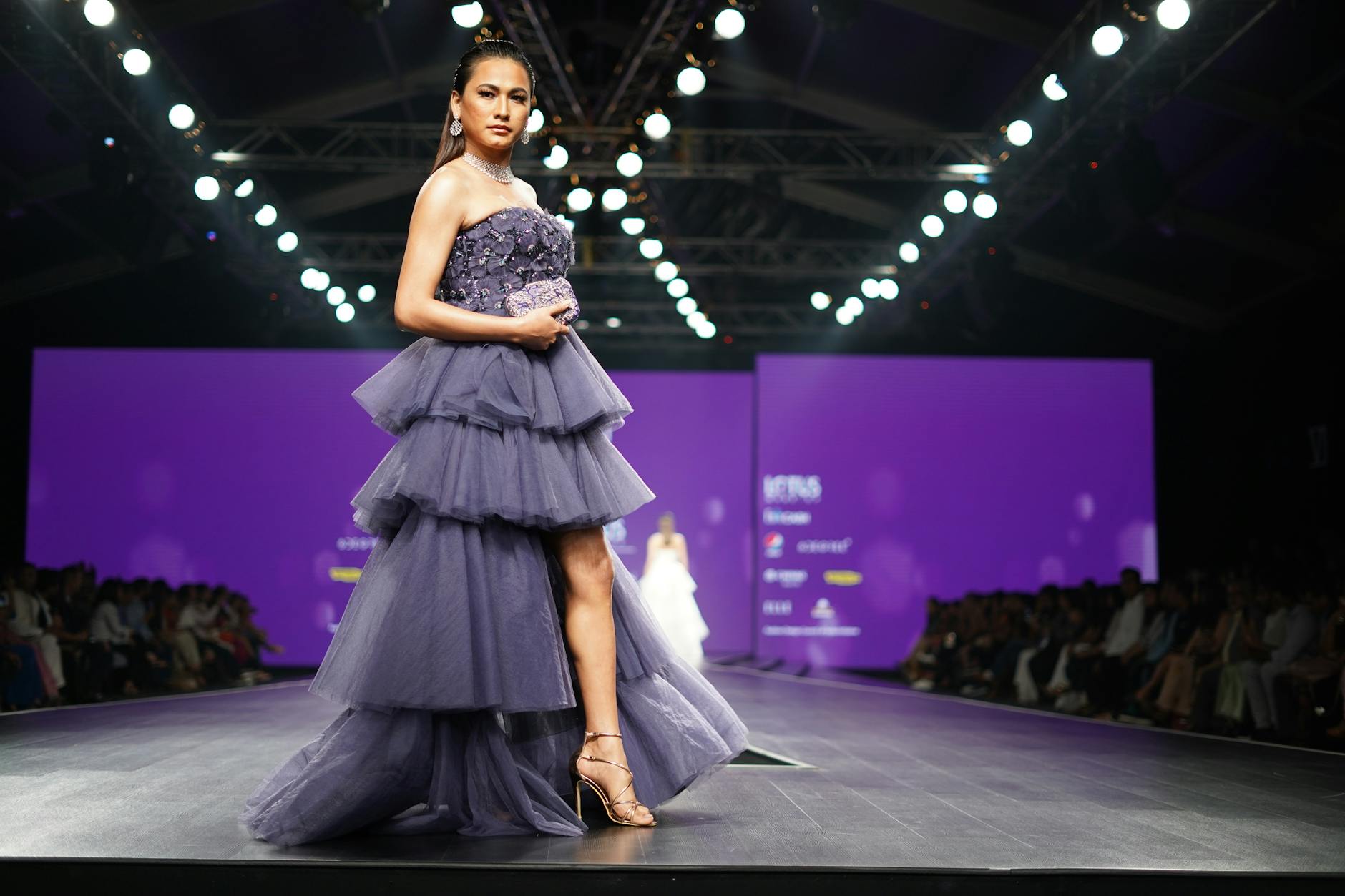 A model poses elegantly in a layered dress on a brightly lit fashion runway stage.