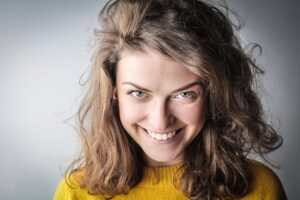 Close-up portrait of a smiling woman in a yellow sweater and casual hair, exuding happiness against a gray background.