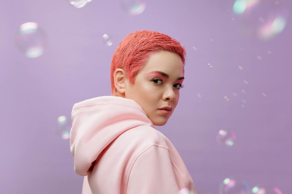 Vibrant studio portrait of a woman with pink hair wearing a hoodie against a lilac background.
