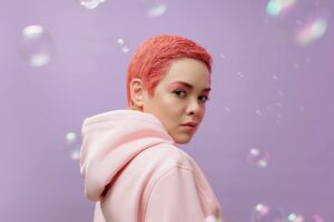 Vibrant studio portrait of a woman with pink hair wearing a hoodie against a lilac background.