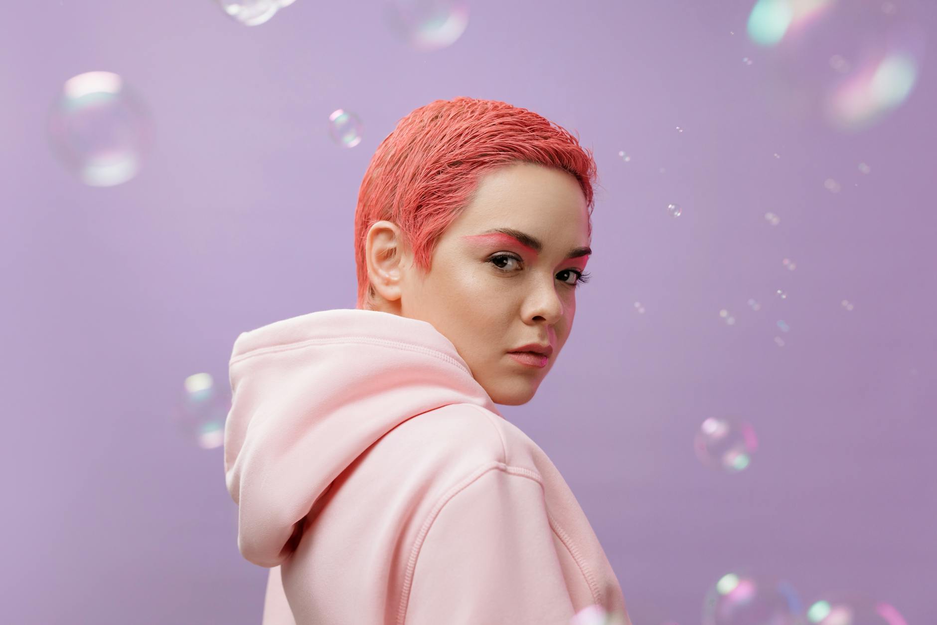 Vibrant studio portrait of a woman with pink hair wearing a hoodie against a lilac background.