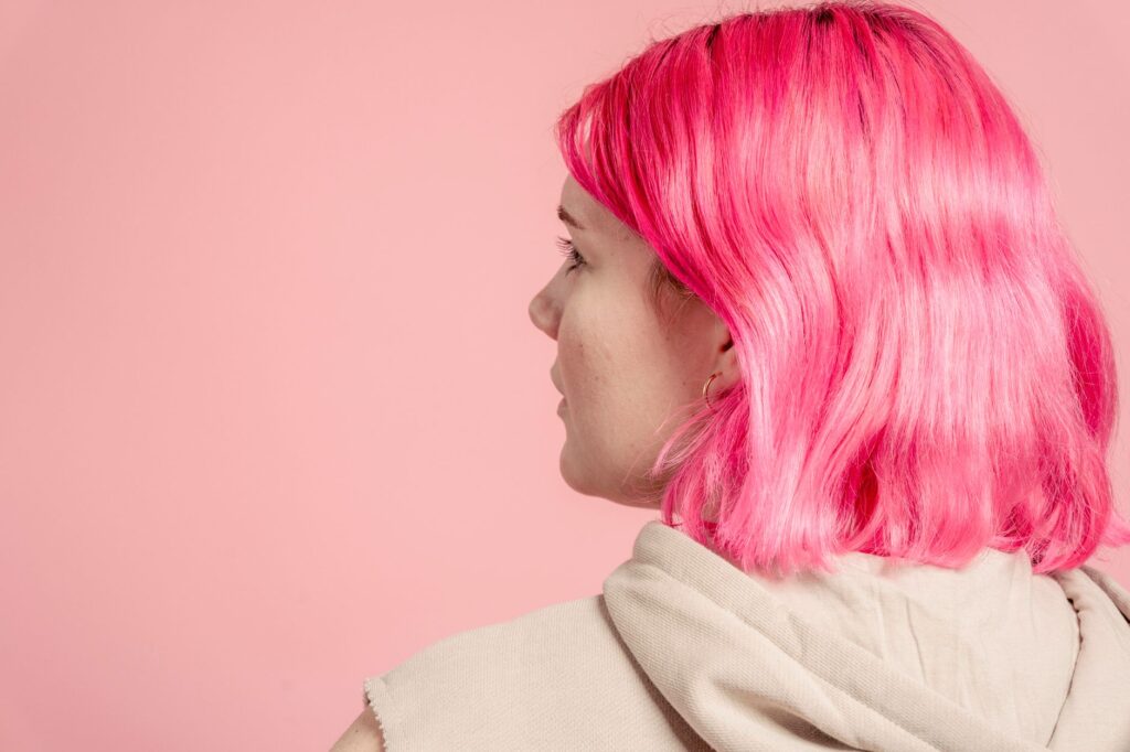 Back view of anonymous young female with dyed hair in casual outfit standing on pink background in modern light studio