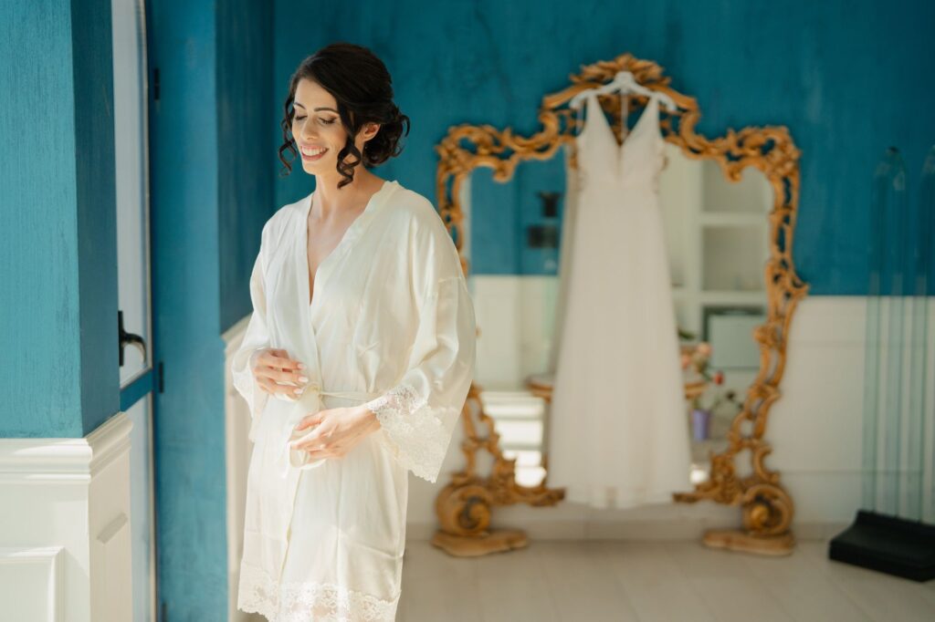 A bride in a robe smiles while preparing for her wedding in a stylish blue room with a gold-framed mirror.