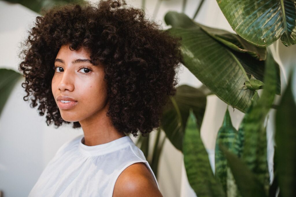 Side view of smiling African American female with Afro hairstyle standing near green plants and looking at camera